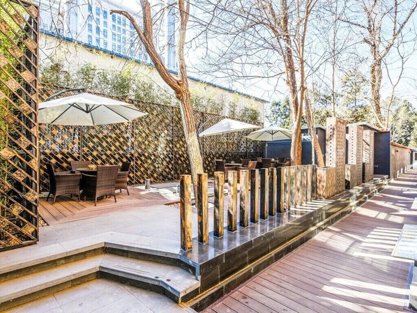 An outdoor patio area with wooden flooring, a seating arrangement with chairs and tables, and umbrellas providing shade.