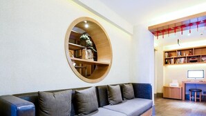 A modern living room with a grey sofa, a wooden circular shelf with plants, and a desk area with a computer and books. - Qiuguo Hotel Beijing by Bayluxury (Beijing)