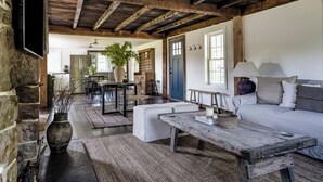 A rustic interior with exposed wooden beams, a stone wall, a dining area with a table and chairs, a sofa, and a large window. - The Farmhouse Stay (Haddam)