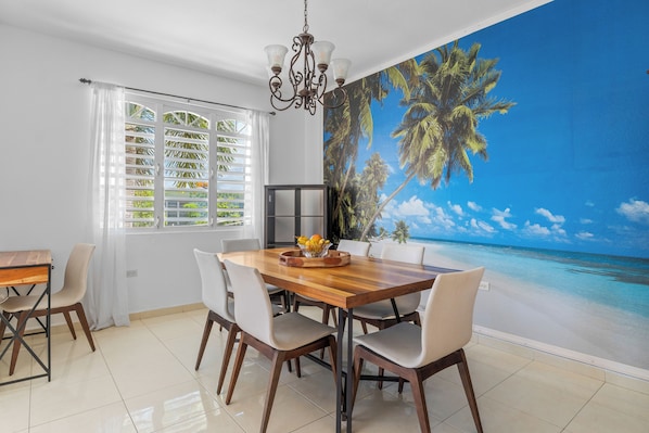 A dining area with a wooden table and chairs, a chandelier, and a large mural of a beach scene.