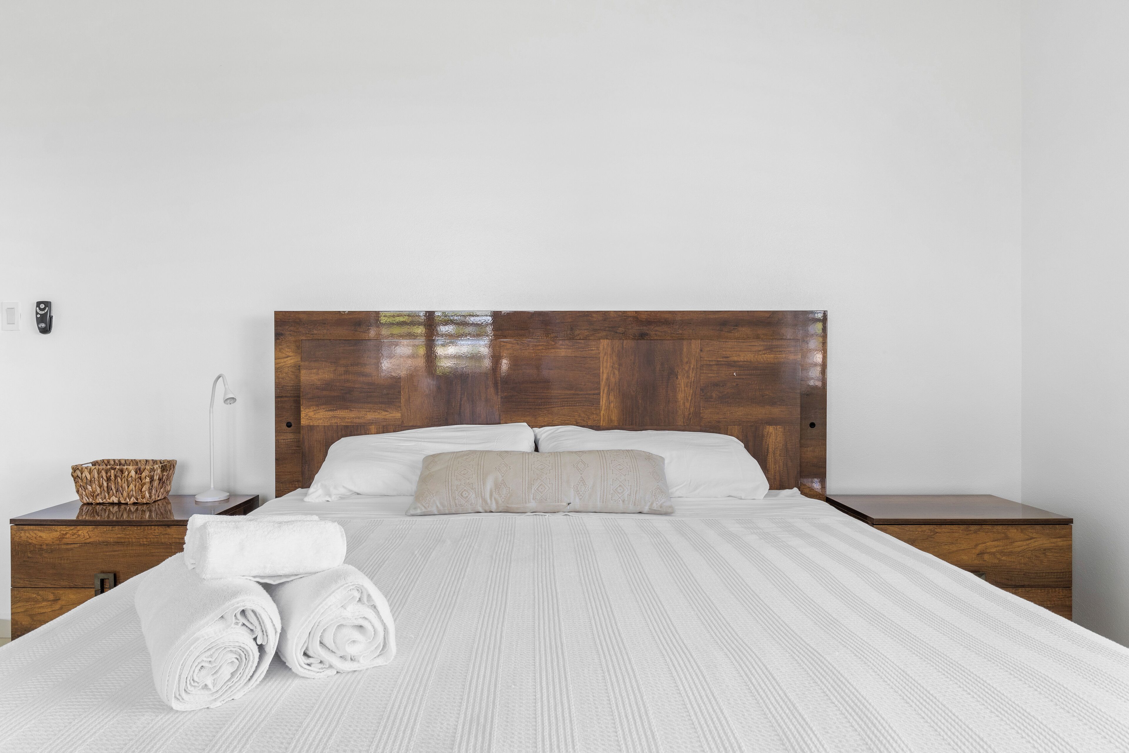 Un letto rifatto con lenzuola bianche e una testiera in legno, una lampada da comodino e un piccolo cesto di vimini su un tavolino di legno.