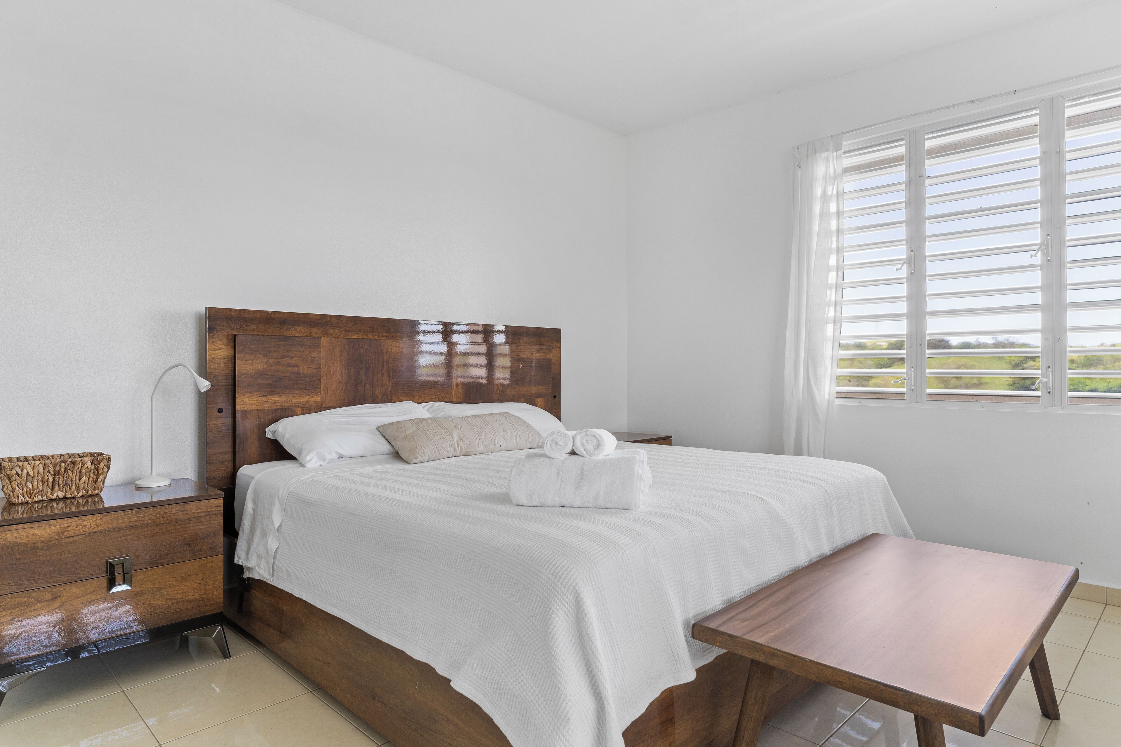 A neatly made bed with white linens, a wooden headboard, a bedside table with a lamp, and a view through a window with horizontal blinds.