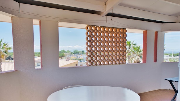 A room with a round table and chairs, a window with a view of buildings and palm trees, and a wall with a circular pattern. - Sea view cool apartment with a balcony (Le Barcarès)