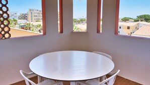 A room with a round table and white chairs, large windows with vertical supports, and a view of buildings and trees outside. - Sea view cool apartment with a balcony (Le Barcarès)