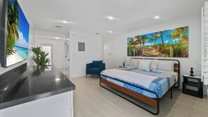 Ein modernes Schlafzimmer mit einem Bett, einem blauen Sessel, einem Fernseher und einem Bild mit Strandmotiv.