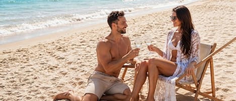 Two people sitting on a beach chair facing the ocean, holding drinks.