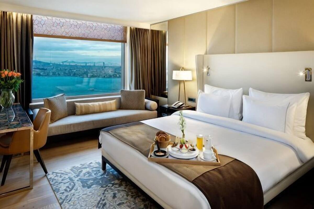 A hotel room with a bed, a desk, a chair, a view of a cityscape, and a nightstand with a lamp.