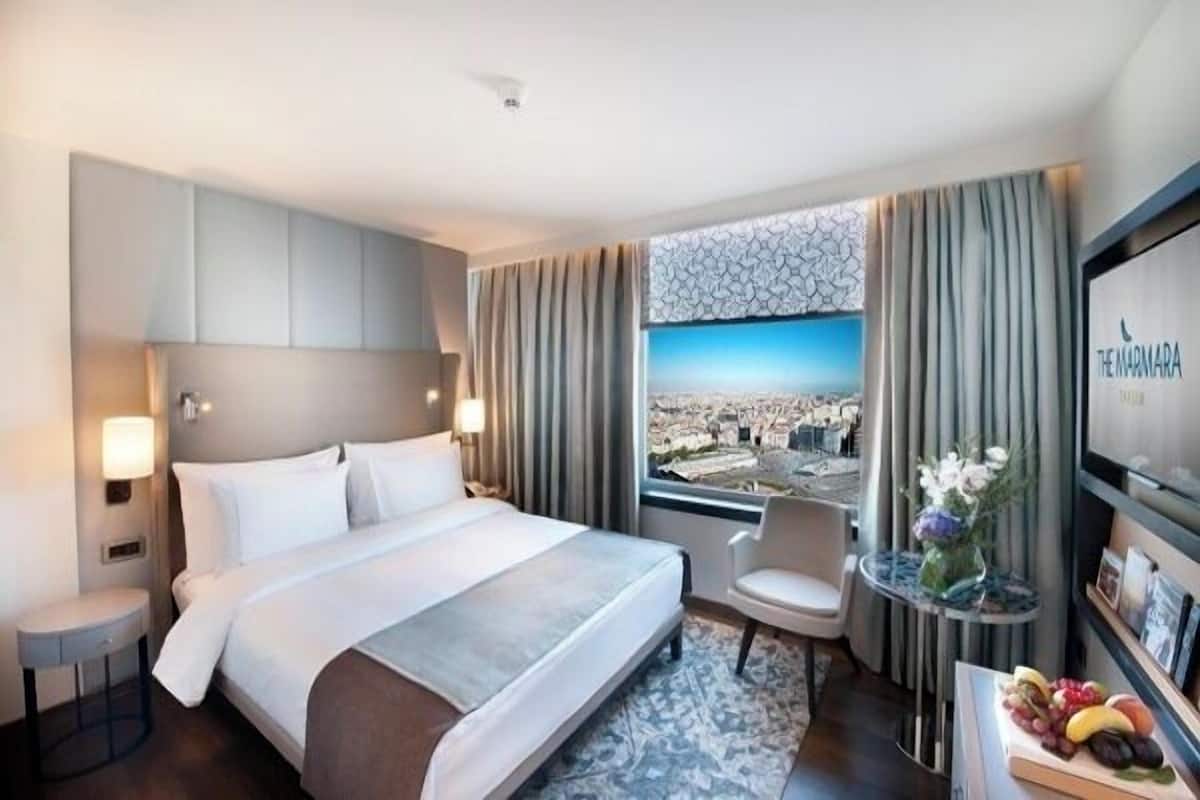 A hotel room with a large bed, a view of the city, a small table with a vase of flowers, and a fruit display.
