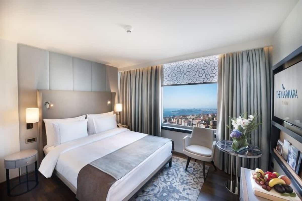 A hotel room with a bed, a chair, a small table with fruit, a vase with flowers, and a city view through the window.
