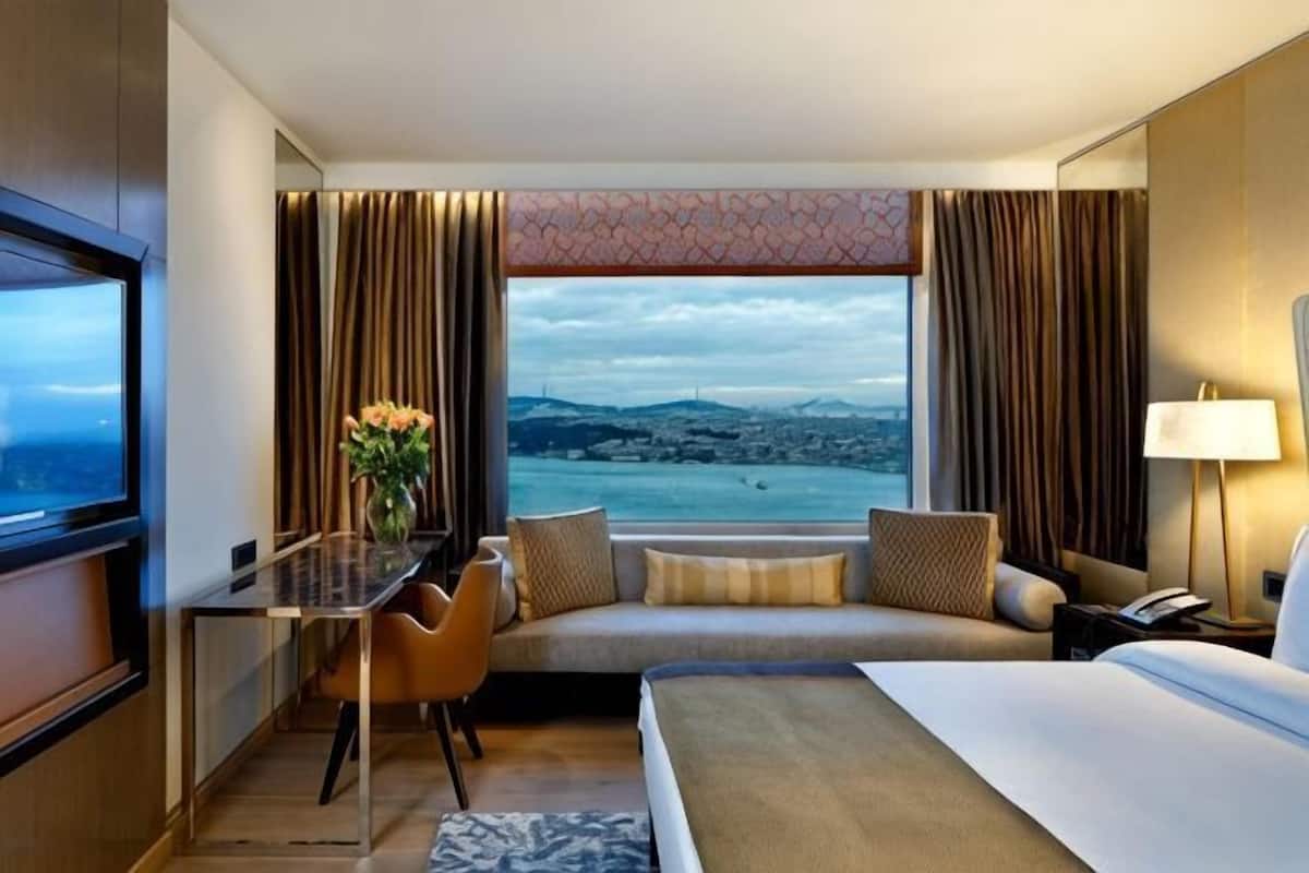 A hotel room with a bed, a desk, a chair, a sofa, a TV, and a view of the city.