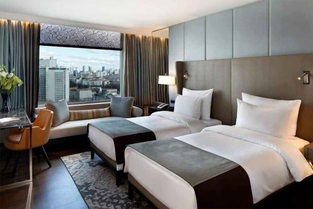 A hotel room with two beds, a desk, a chair, and a view of the city.