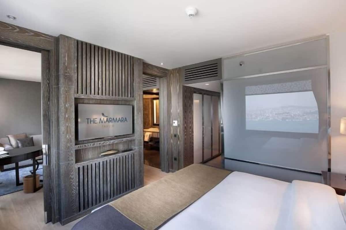 A modern hotel room with a large bed, a flat-screen TV mounted on a wooden panel, a seating area with a table and chairs, and a cityscape view projected on the wall.