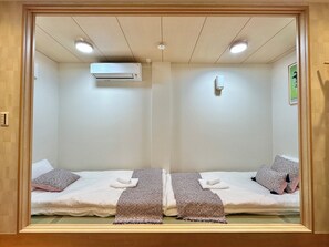 A room with two beds, each with a pillow and a blanket, an air conditioning unit, a wall-mounted light, and a framed picture on the wall.