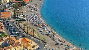 Un poble costaner amb una platja, un castell històric sobre un turó i una zona d'aparcament amb cotxes.