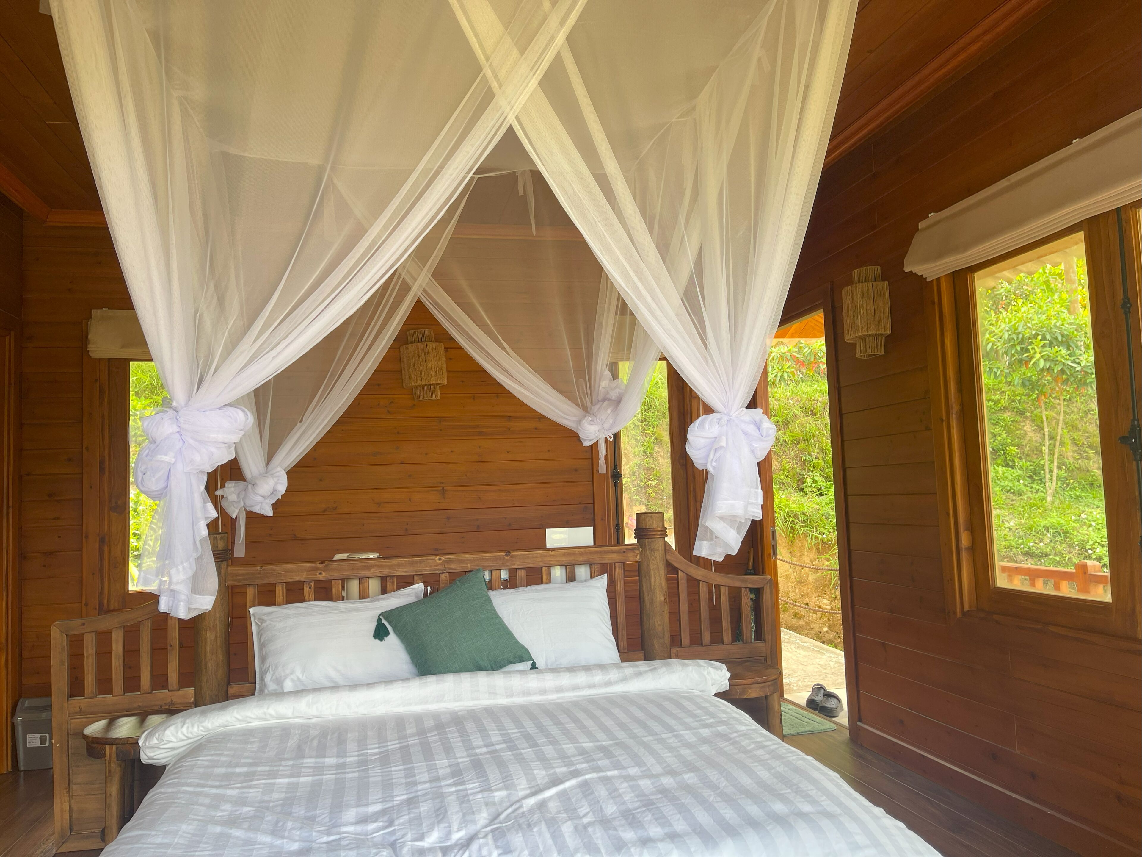 A wooden cabin bedroom with a bed, white curtains, and a view of greenery outside.