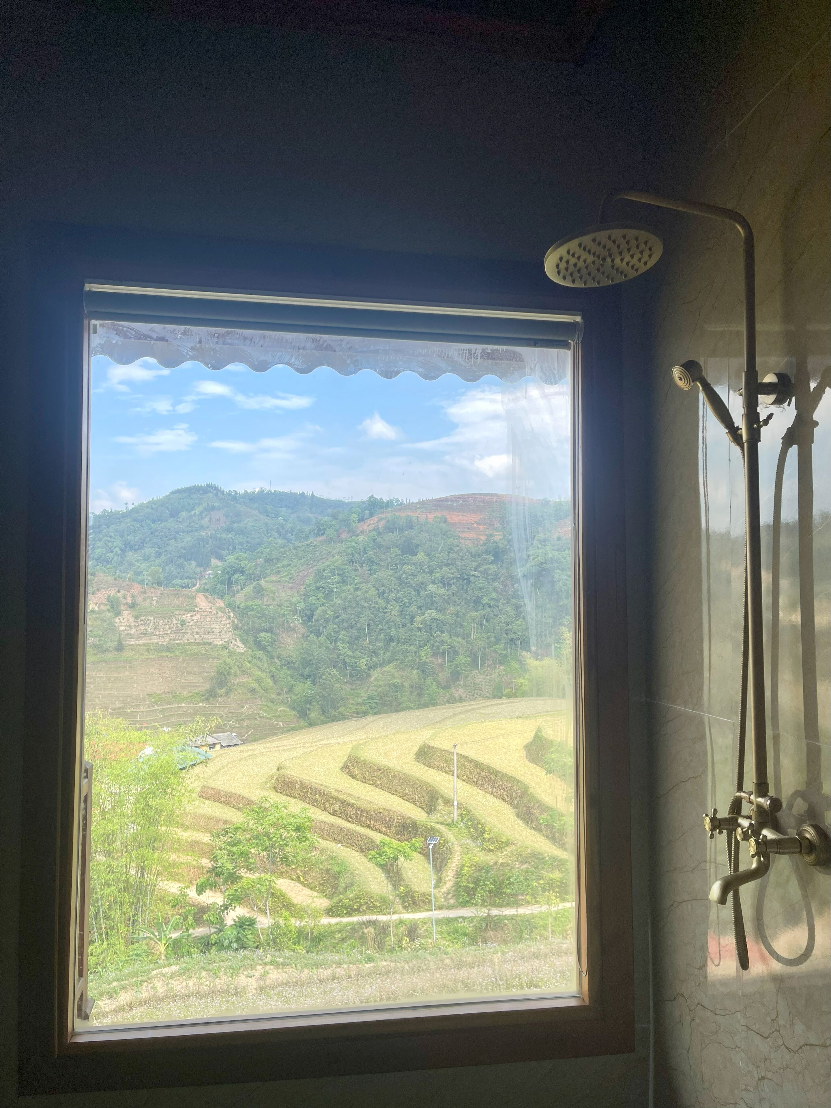 Un baño con ducha, una ventana con vistas a campos escalonados y colinas.