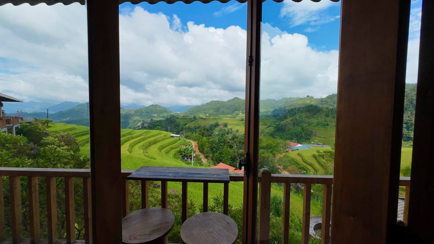 A view from a balcony with a wooden railing, overlooking terraced fields and distant hills.