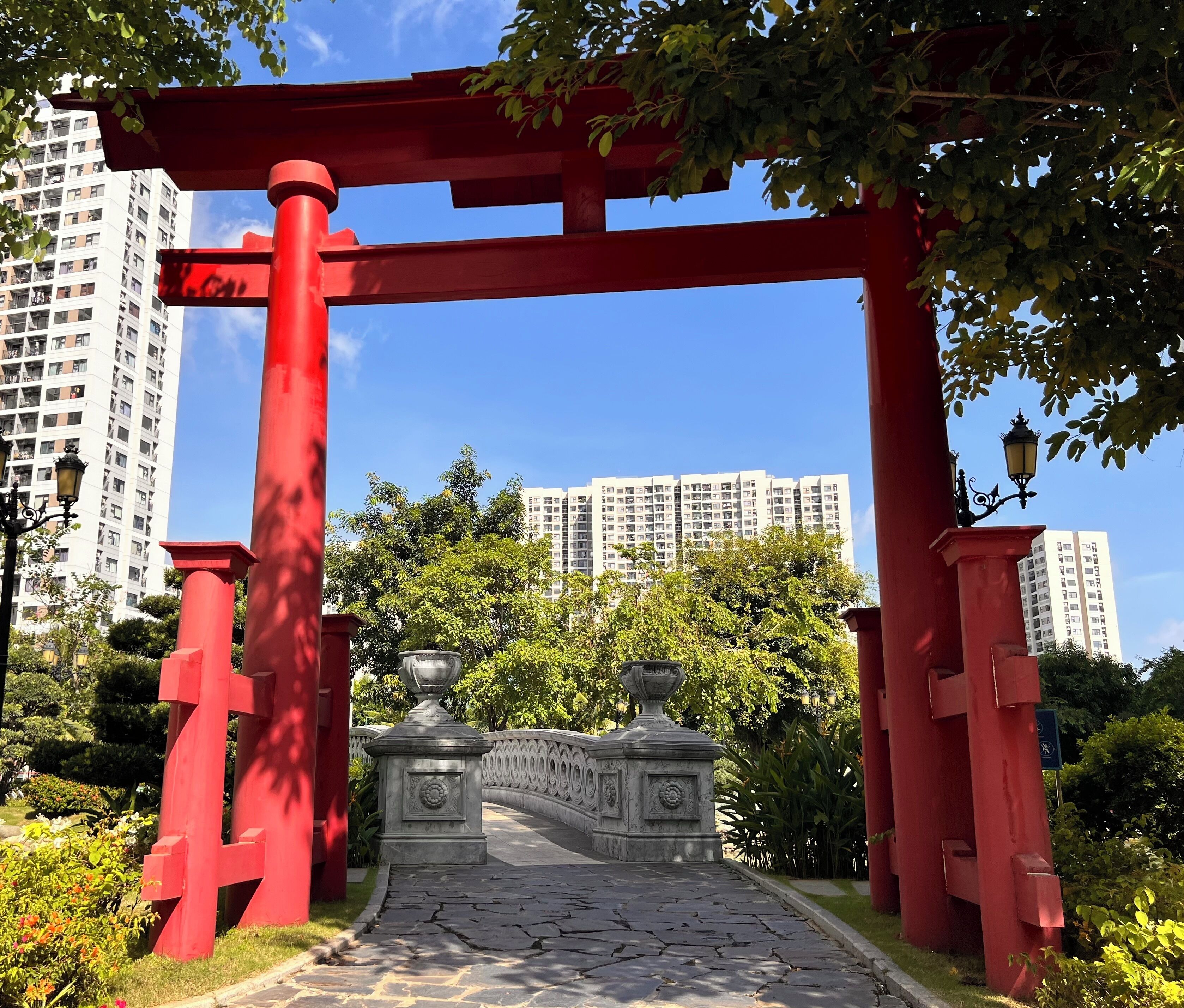 A red torii gate leading to a stone pathway in a park with greenery and high-rise buildings in the background.