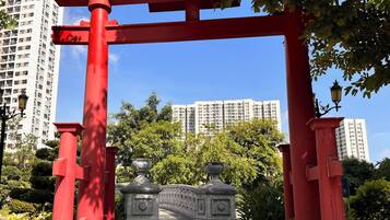 A red torii gate leading to a stone pathway in a park with greenery and high-rise buildings in the background.