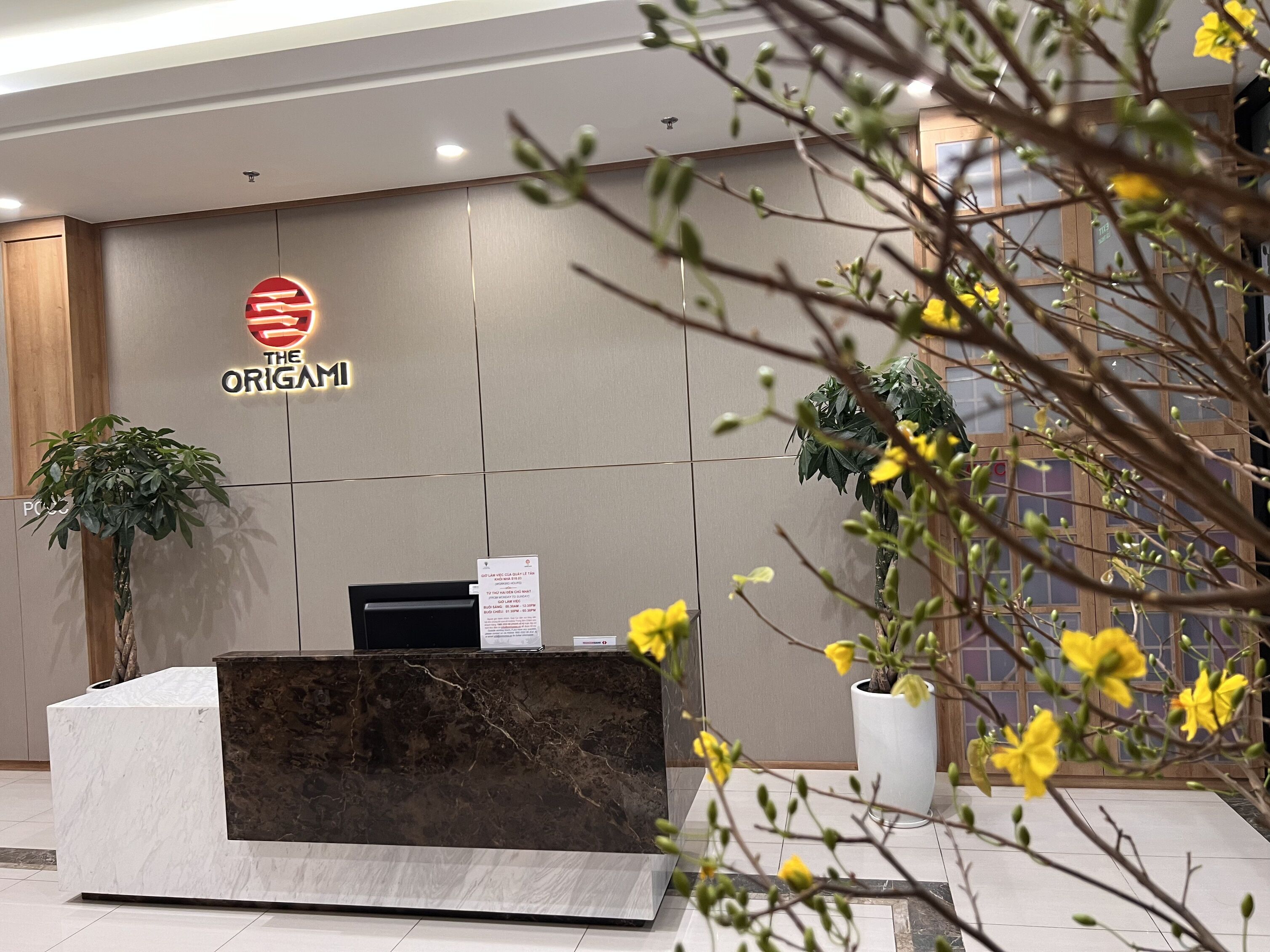 A reception desk with a marble top and a dark marble base, a sign reading "The Origami", and a potted plant to the left.