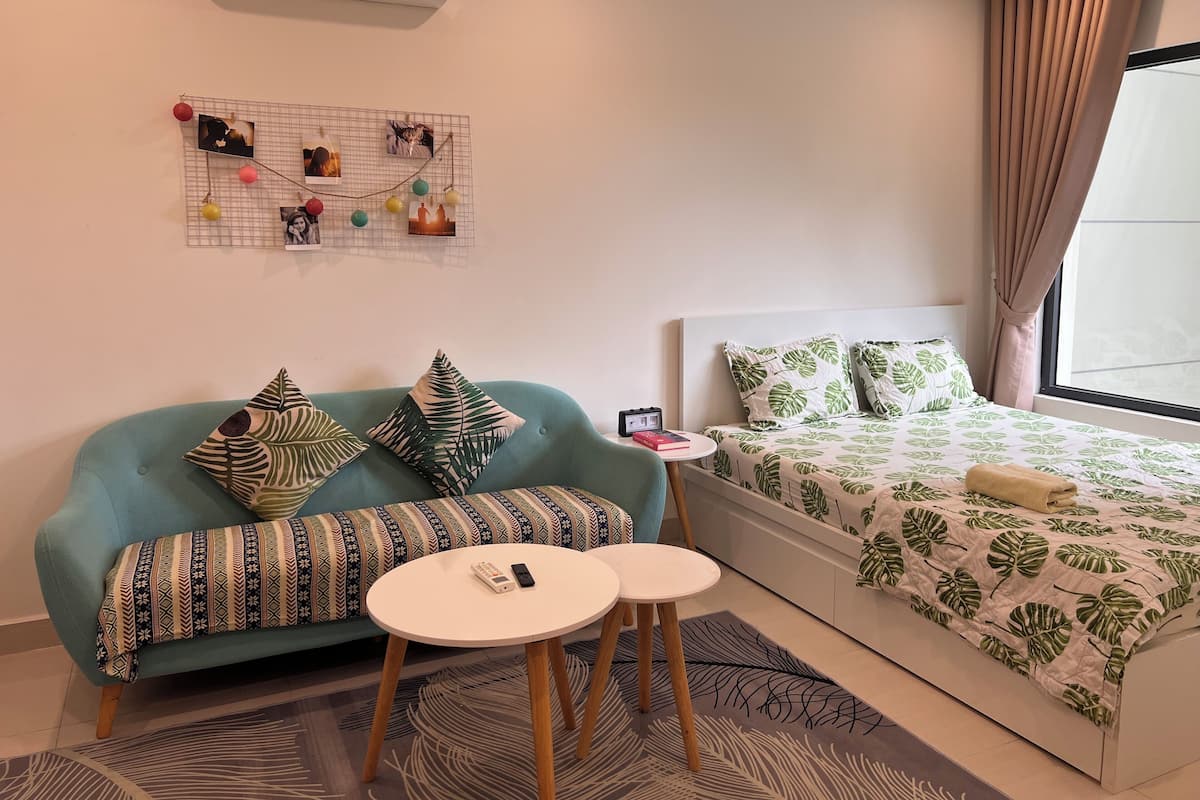 Une chambre d’hôtel moderne avec un canapé de couleur turquoise, un lit recouvert d’une couvre-lit à motifs de feuilles, une petite table de chevet avec une horloge et un présentoir photo fixé au mur.