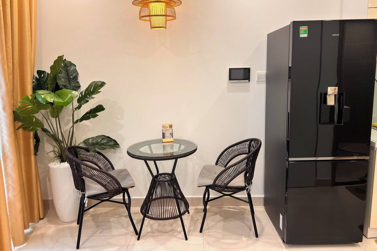 Un espace de vie compact comprenant un réfrigérateur noir, deux fauteuils en osier, une petite table en verre, une plante en pot et un luminaire suspendu moderne.