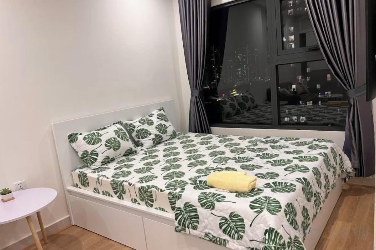 Una cama con colcha estampada de hojas, una mesita de noche y una ventana con cortinas.