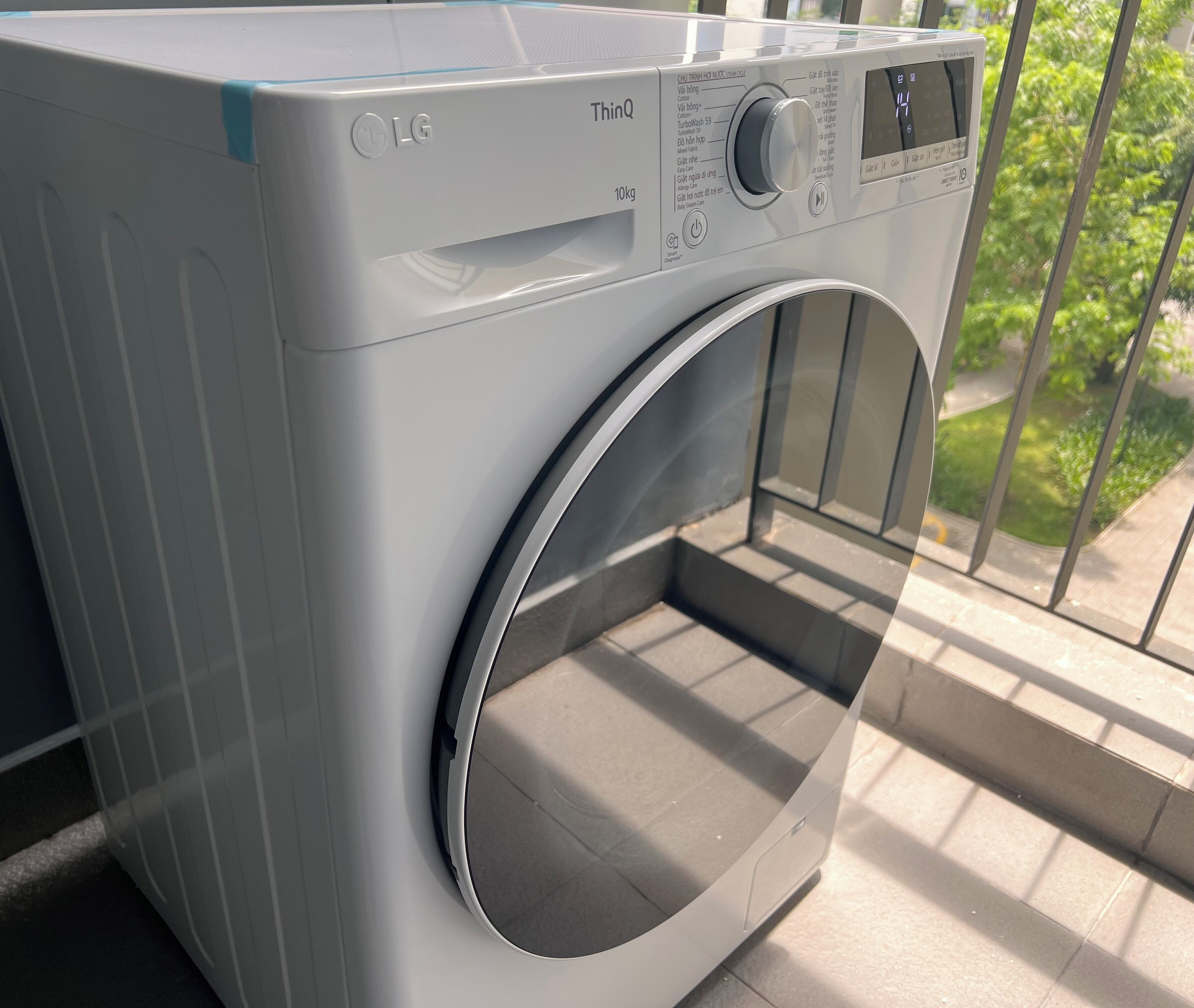 A front view of a white LG ThinQ washing machine with a digital display and control panel, placed on a balcony with a view of greenery.