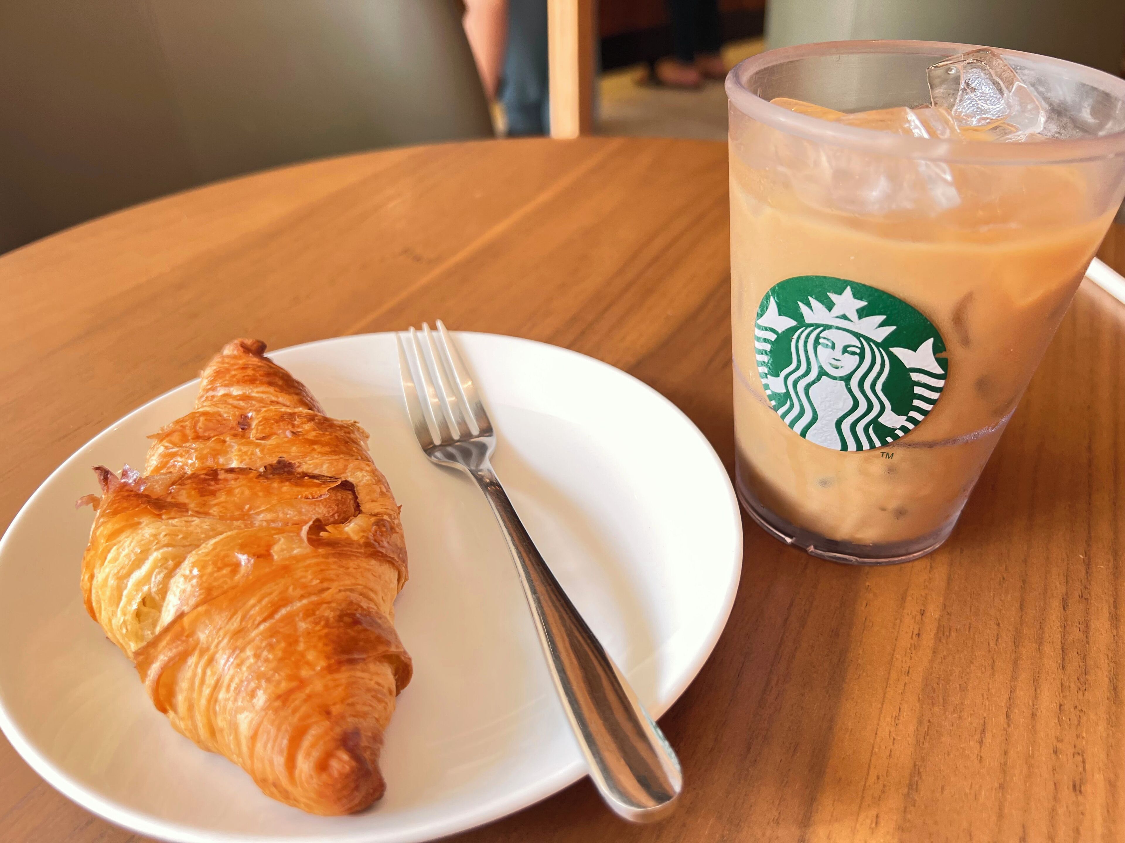 Un croissant sur une assiette blanche, avec une fourchette à côté, et un gobelet de café glacé de Starbucks avec une paille.