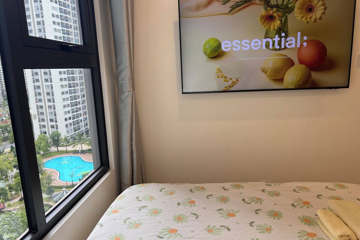 Une chambre d’hôtel avec un lit, une fenêtre donnant sur une piscine et des immeubles de grande hauteur, et un téléviseur fixé au mur qui affiche une image florale.