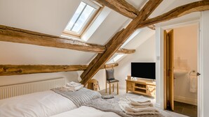 A bedroom with a sloped ceiling, wooden beams, a bed, a TV, and a bathroom visible through an open door.