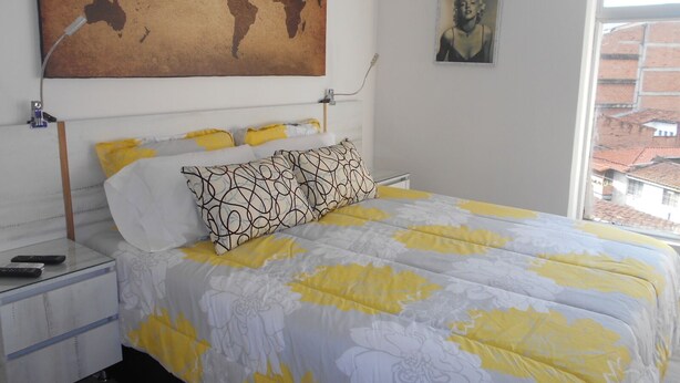 Un dormitorio con una cama, una mesita de noche, un mapa mundial en la pared y una ventana con vistas a edificios.