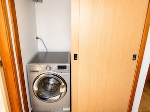 A compact, stainless steel front-loading washing machine in a small laundry room with a wooden door and a white wall.