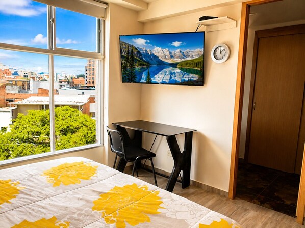 Habitación de hotel con cama, escritorio, silla, televisor en la pared y vista a un paisaje urbano a través de un ventanal grande.