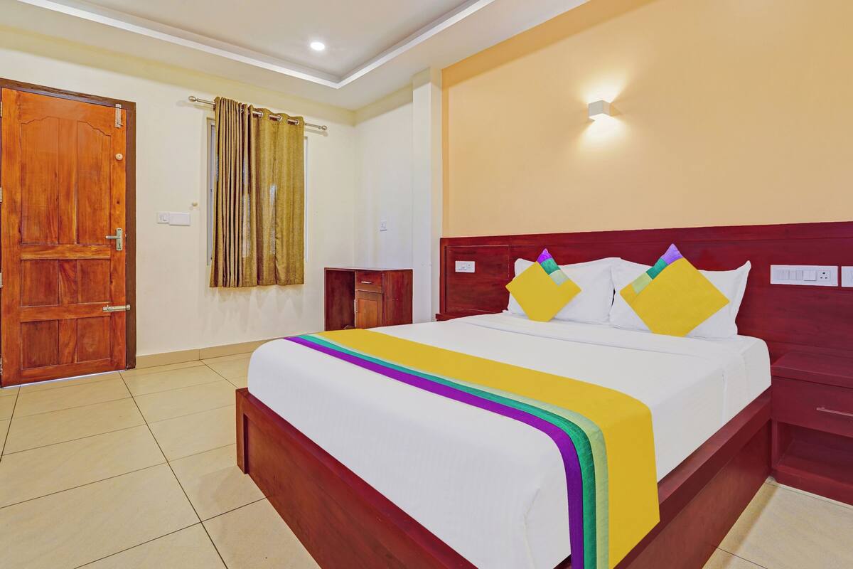 A hotel room with a wooden door, a bed with a colorful bedspread, a nightstand, and a red headboard.