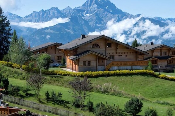 Edificio de estilo chalete con techo inclinado, rodeado de un césped cuidado y con montañas al fondo.