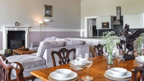 Een eetgedeelte met een houten tafel voor vier personen, omringd door beklede stoelen. Achtergrond toont een open haard met een manteltas en gedeeltelijk een keuken door een open doorgang.