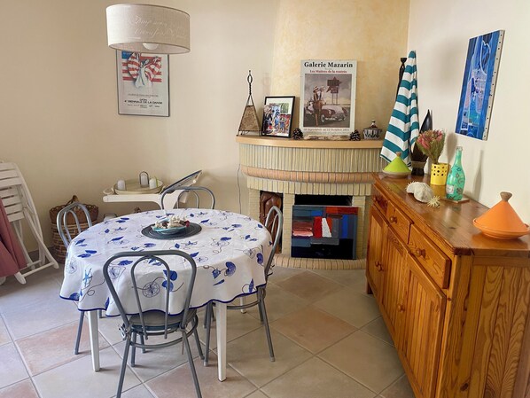Uma sala de jantar com uma mesa redonda coberta por um tecido floral azul, rodeada por cadeiras de metal. Um armário de madeira com um tecido verde e branco fica à direita.