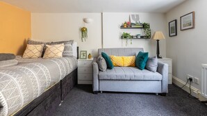 A bedroom with a bed, a grey sofa, a bedside table, a lamp, and wall-mounted shelves with plants. - Cozy Flat in the Heart of Camden (Greater London)
