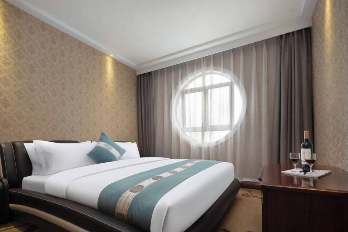 A hotel room with a bed, a round window, a nightstand with a bottle and glasses, and a carpeted floor.