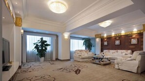 Un salón espacioso con un amplio sofá, una mesa de centro y un televisor. Hay dos ventanas grandes con cortinas, un candelabro y un panel decorativo en la pared.