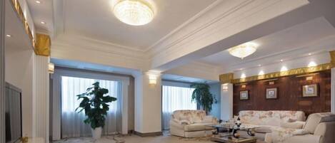 Un salon spacieux comprenant un grand canapé, une table basse et un téléviseur. Il y a deux grandes fenêtres avec des rideaux, un lustre et un panneau décoratif au mur.