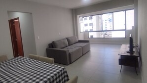 A living room with a sofa, a dining table with a checkered tablecloth, and a large window with a view of buildings. - Cozy 2 Bedroom Apartment in the center of Camboriú, 200 meters from the beach (Centro Balneário Camboriú)