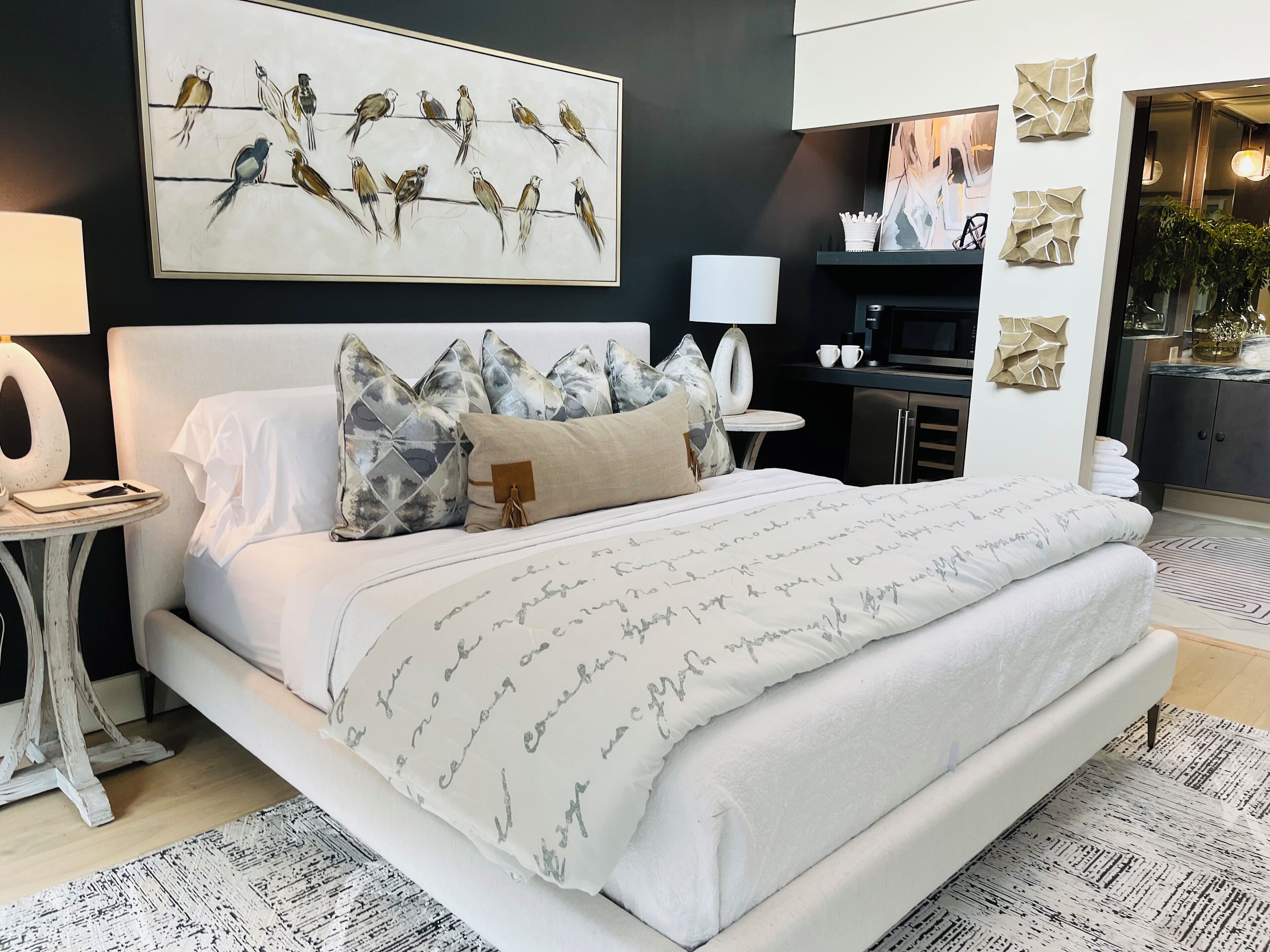 Une chambre à coucher avec un grand lit, une table de chevet, une commode, une lampe fixée au mur et un tableau représentant des oiseaux.