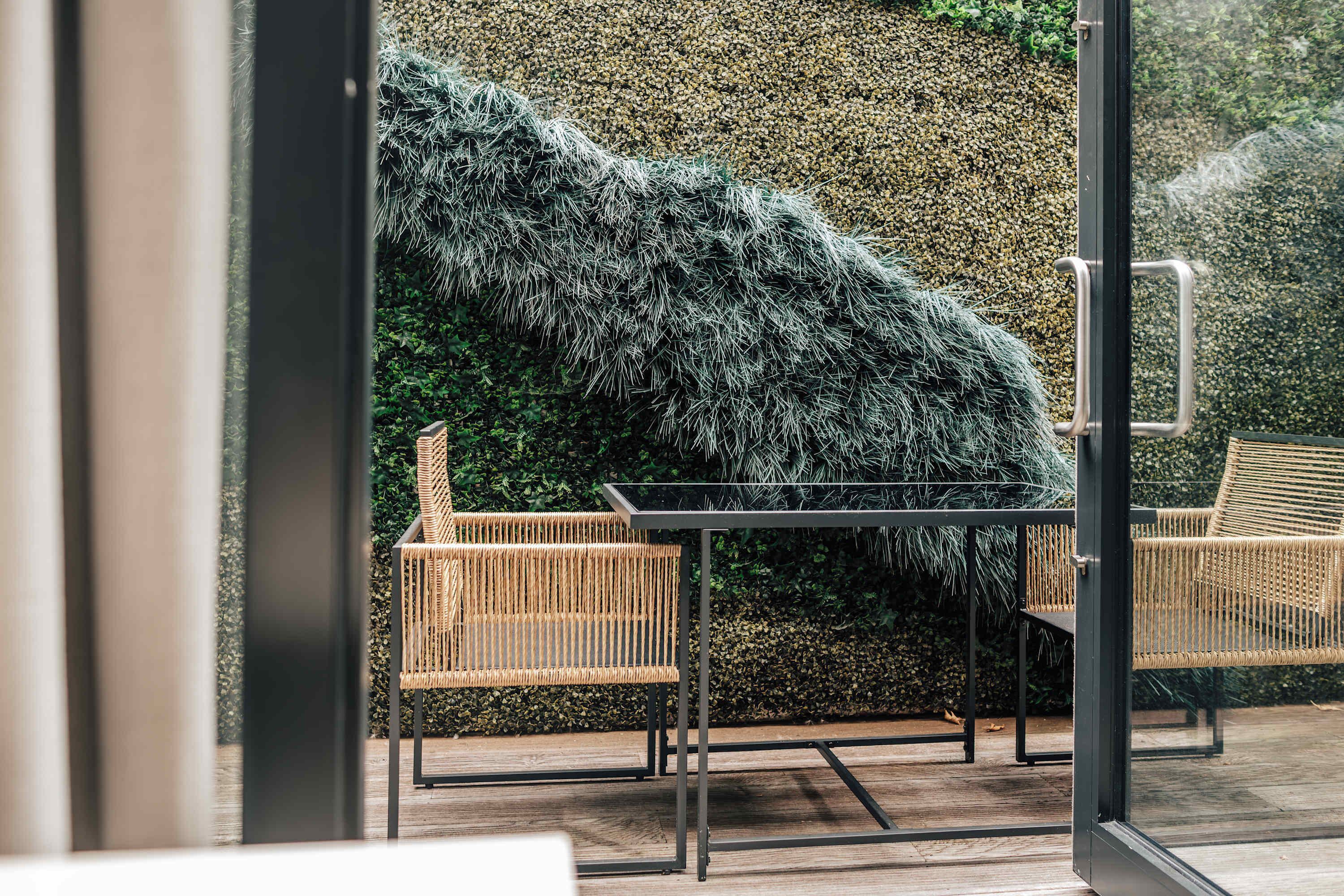 Ein moderner Außenbereich mit Korbstühlen und einem Glastisch, vor einer strukturierten Wand und einer Holzterrasse.