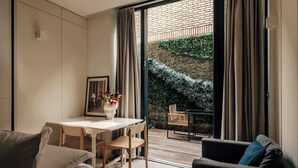 A modern living room with a dining area, a sofa, a coffee table, and a view of a brick wall and greenery outside. - The Westlight: Bright LG Studio in White City (London)