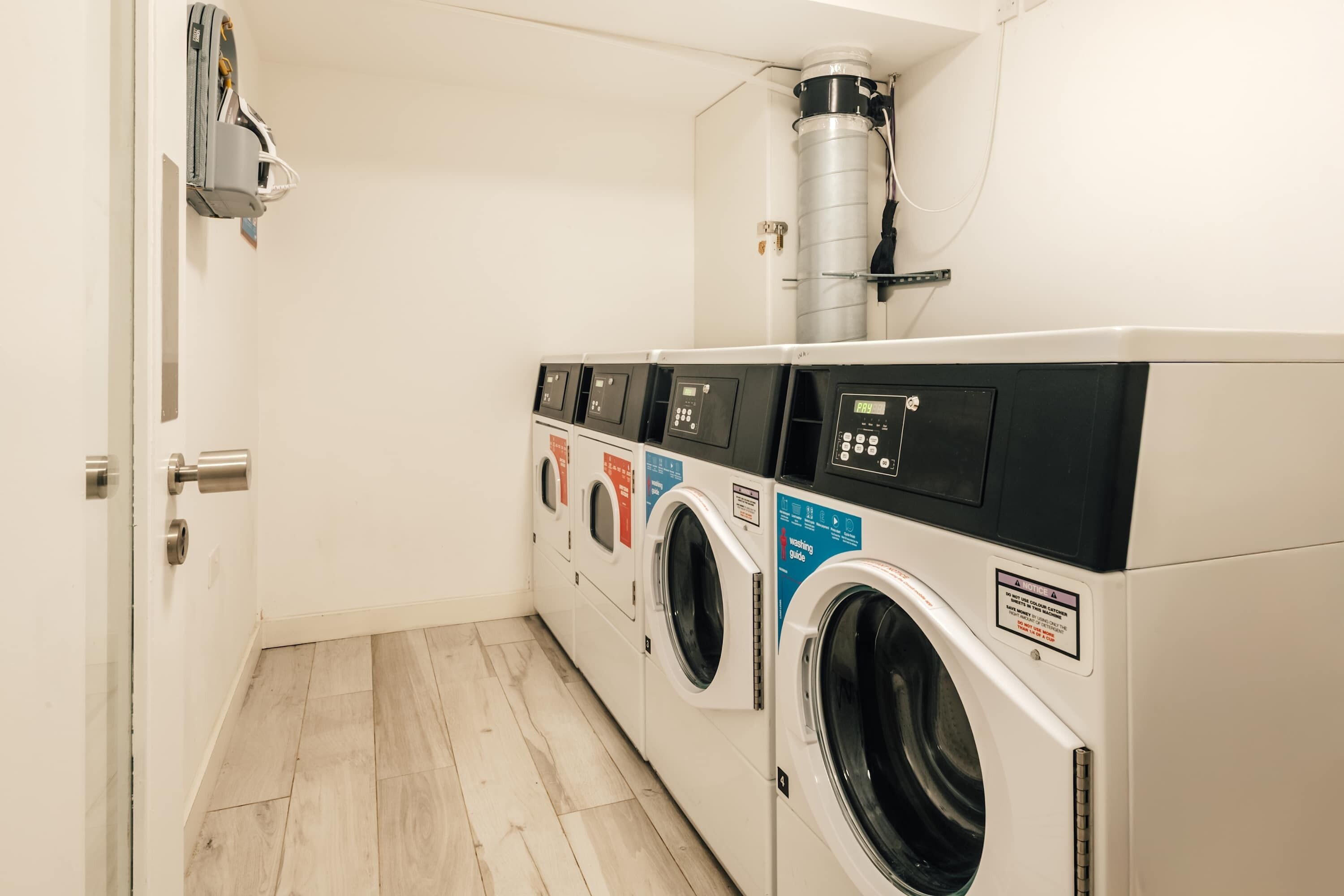 A laundry room with multiple washing machines and dryers, a ventilation system, and a door.
