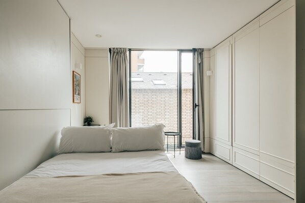 Une chambre à coucher au style minimaliste, avec un grand lit, une petite table de chevet et une vue sur un bâtiment en briques, visible à travers une porte coulissante.