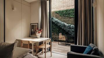 A modern living room with a dining area, a sofa, a coffee table, and a view of a brick wall and greenery outside.