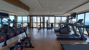 A gym with treadmills, dumbbells, and exercise equipment, large windows with a city view. - COBAR0114 - Edifício Barra Premium (Salvador)